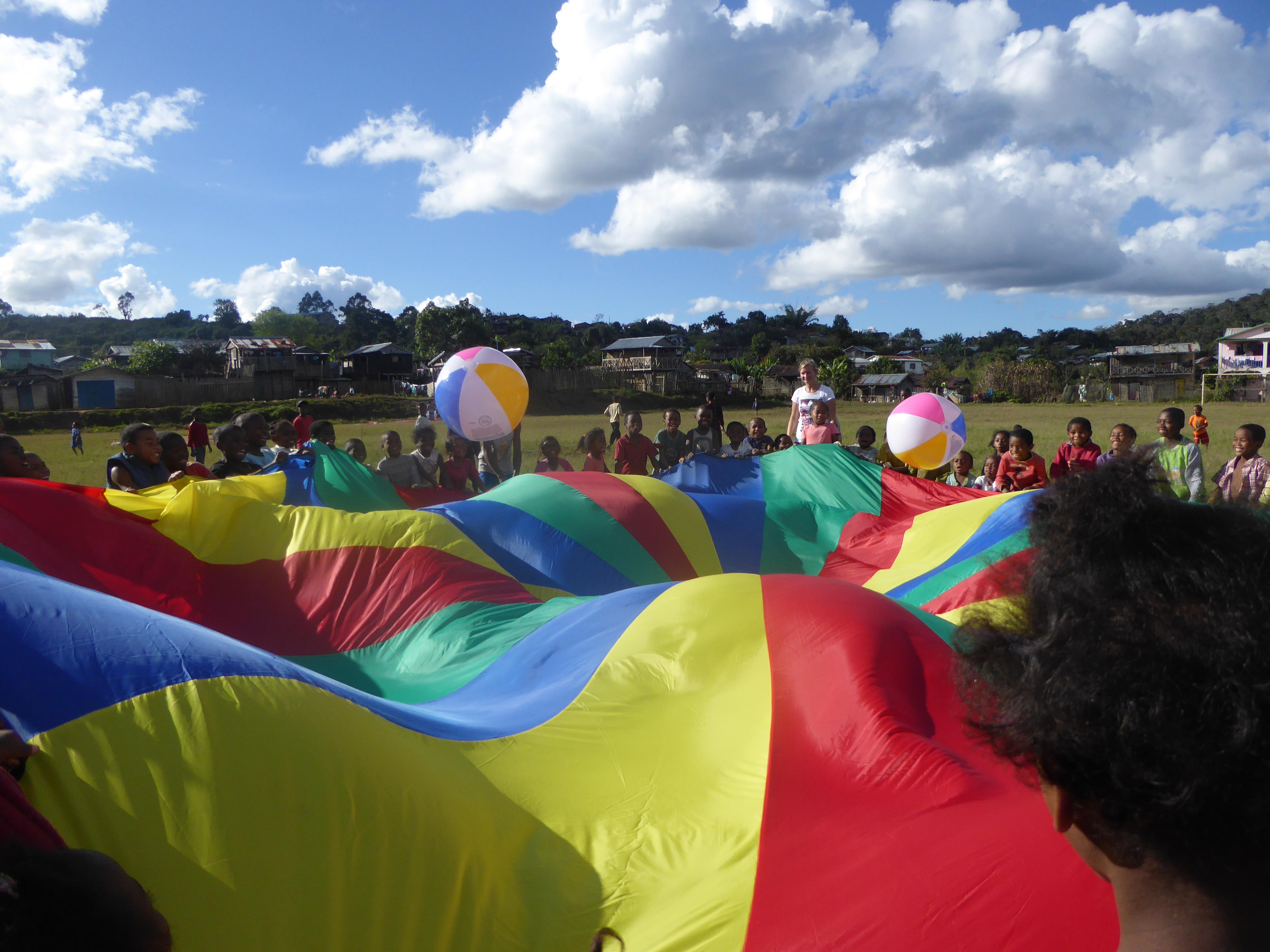 Childcare volunteers play ball games with children using a big parachute outside a school in Madagascar. Projects Abroad volunteers with children in Madagascar are playing outdoor activities with children using a big parachute to develop motor skills in a school.